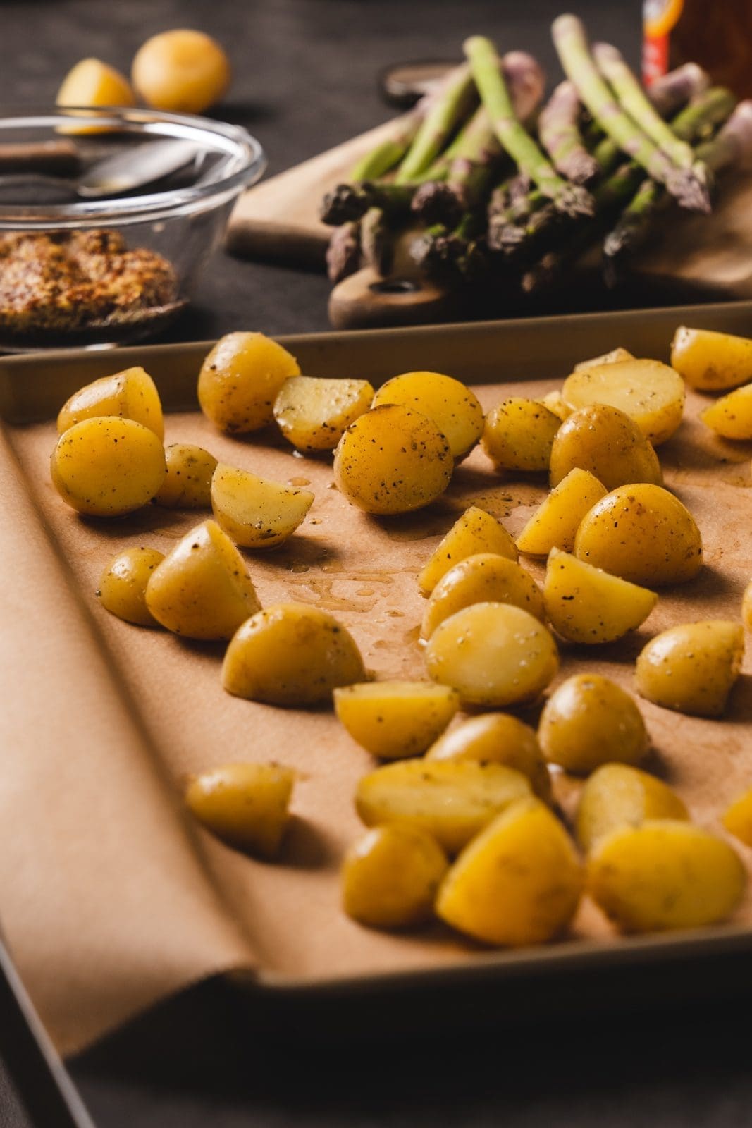 Halbierte Drillinge eingeölt auf dem Backblech. Im Hintergrund grüner Spargel