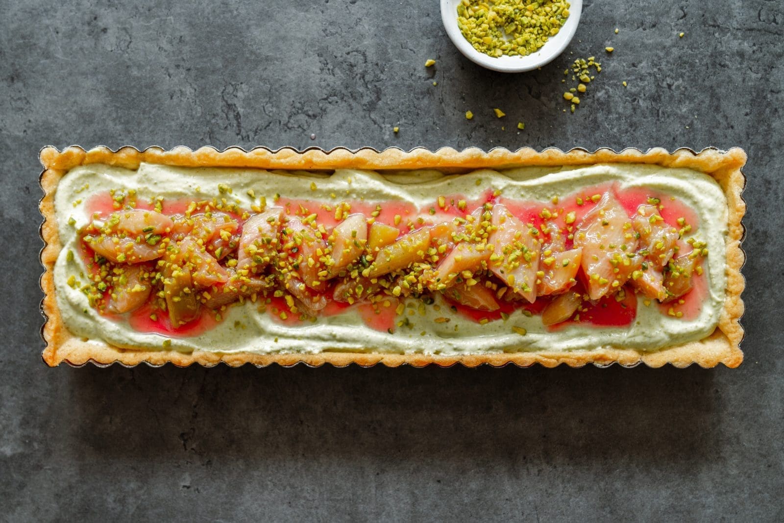 Pistazien-Tarte mit Rhabarber quer im Bild mit gemahlenen Pistazien im Hintergrund