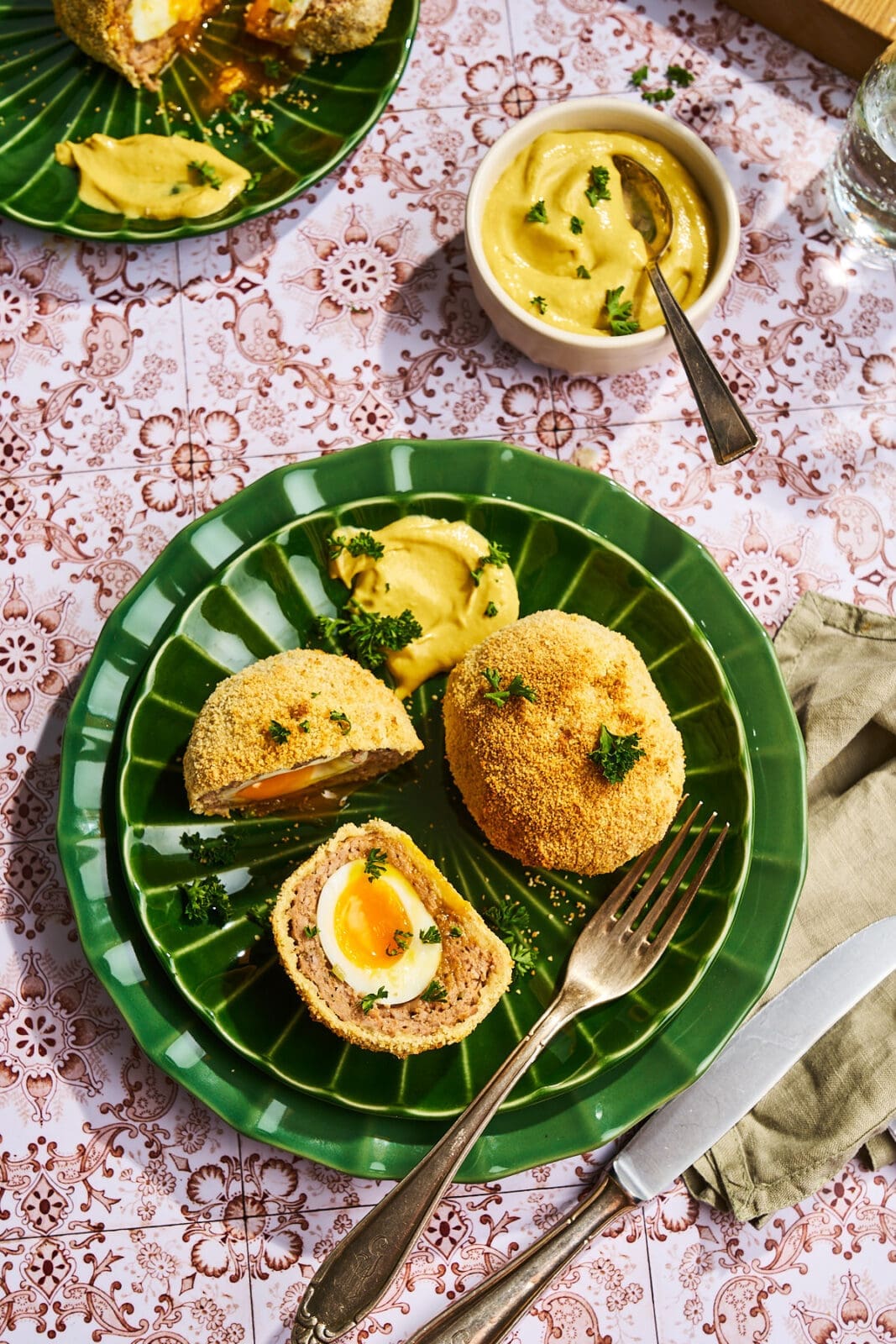 Draufsicht auf eine Servierplatte mit Scotch Eggs, einer Schale Senf und Besteck auf einer gemusterten Tischdecke.