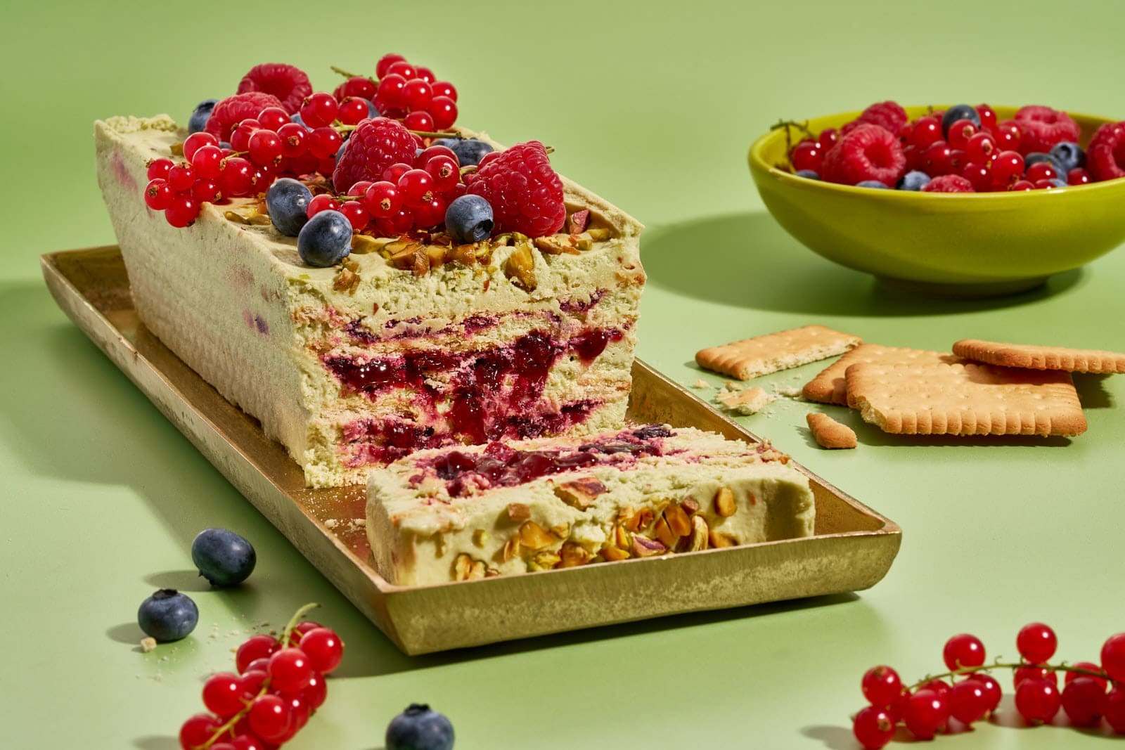 Pistazienkuchen ohne Backen. Cremiger Sommerkuchen mit Waldbeeren aus dem Kühlschrank 4 Pistazienkuchen ohne Backen mit Waldbeeren auf goldenem Tablett, angeschnitten, daneben Butterkekse und eine Schale frische Beeren