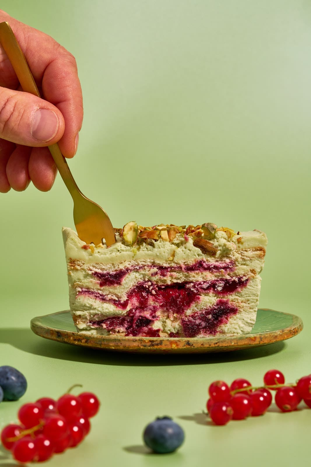 Pistazienkuchen ohne Backen. Cremiger Sommerkuchen mit Waldbeeren aus dem Kühlschrank 5 Goldene Kuchengabel sticht in ein Stück Pistazienkuchen ohne Backen mit Waldbeeren auf grünem Teller