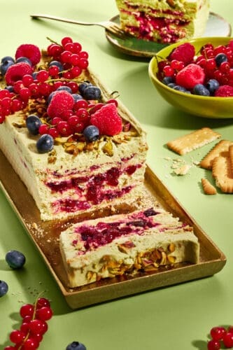 Pistazienkuchen ohne Backen mit Waldbeeren auf goldenem Tablett, daneben Butterkekse und eine Schale mit frischen Beeren