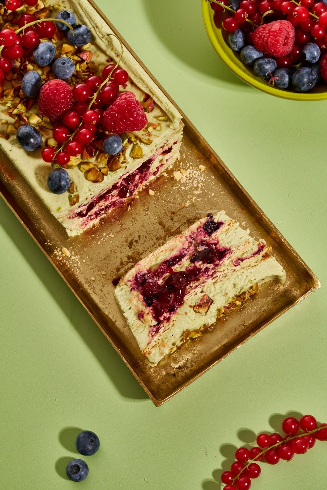 Pistazienkuchen ohne Backen. Cremiger Sommerkuchen mit Waldbeeren aus dem Kühlschrank 9 Pistazienkuchen ohne Backen auf goldenem Tablett, seitliche Ansicht mit sichtbarem Waldbeeren-Kompott zwischen den Cremeschichten