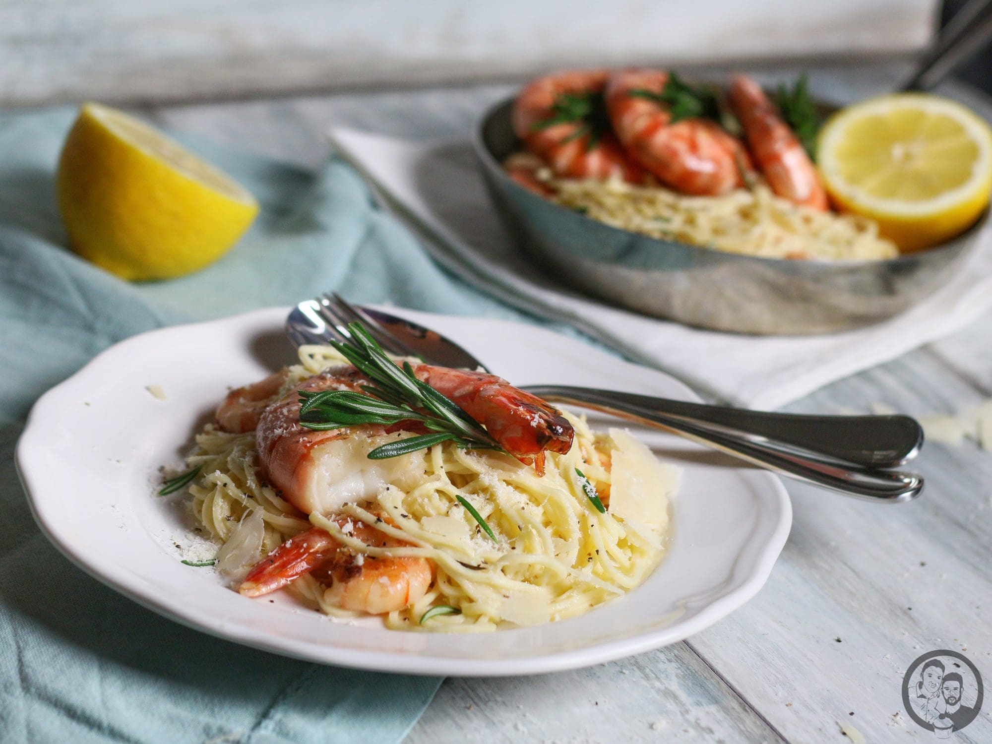 Eine Portion Pasta mit Garnelen und Rosmarin auf einem weißen Teller, im Hintergrund die Pfanne mit den restlichen Garnelen